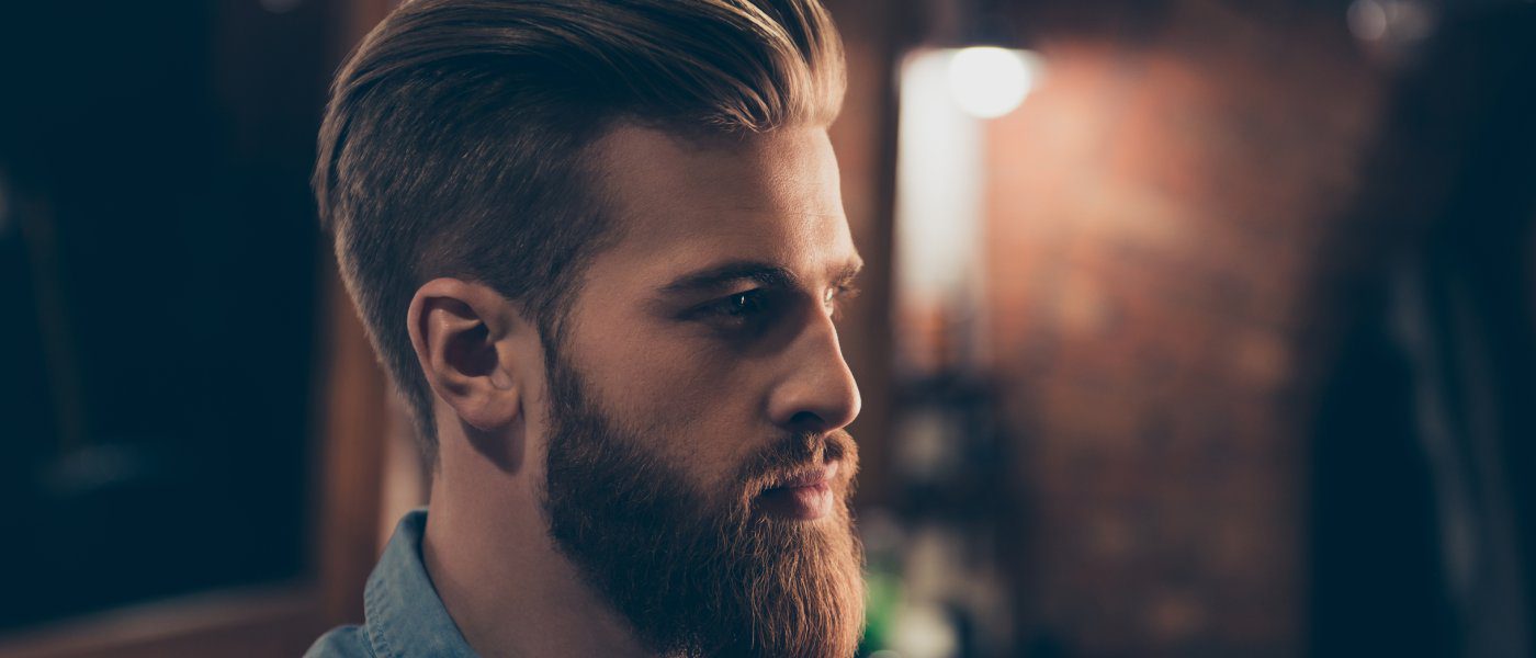 A handsome man standing with his side profile exposed. He has brown hair, nicely groomed and a grown out beard.