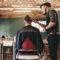 A trendy, industrial styled barbershop with a client in the salon chair. The barber is shaving the sides of his head.