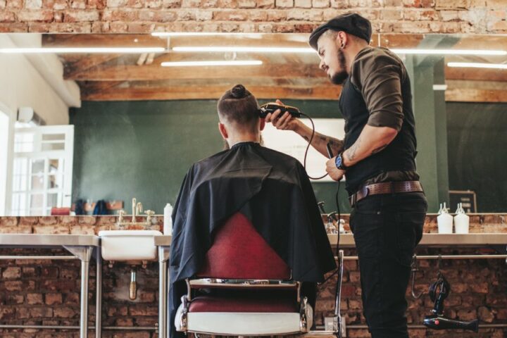 A trendy, industrial styled barbershop with a client in the salon chair. The barber is shaving the sides of his head.