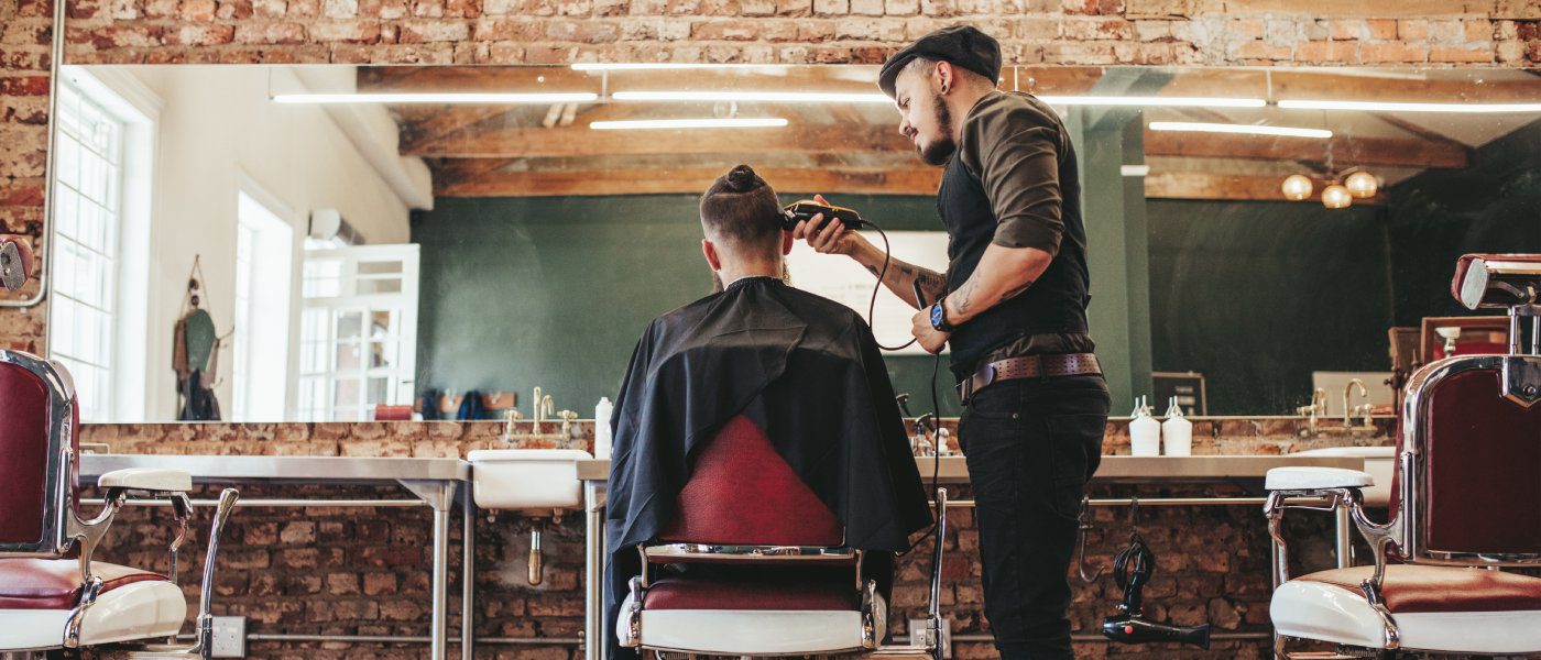 A trendy, industrial styled barbershop with a client in the salon chair. The barber is shaving the sides of his head.