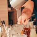 A man standing behind a home bar countertop, straining a drink into a glass. Several liquors and syrups line the counter.