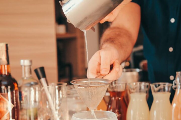 A man standing behind a home bar countertop, straining a drink into a glass. Several liquors and syrups line the counter.