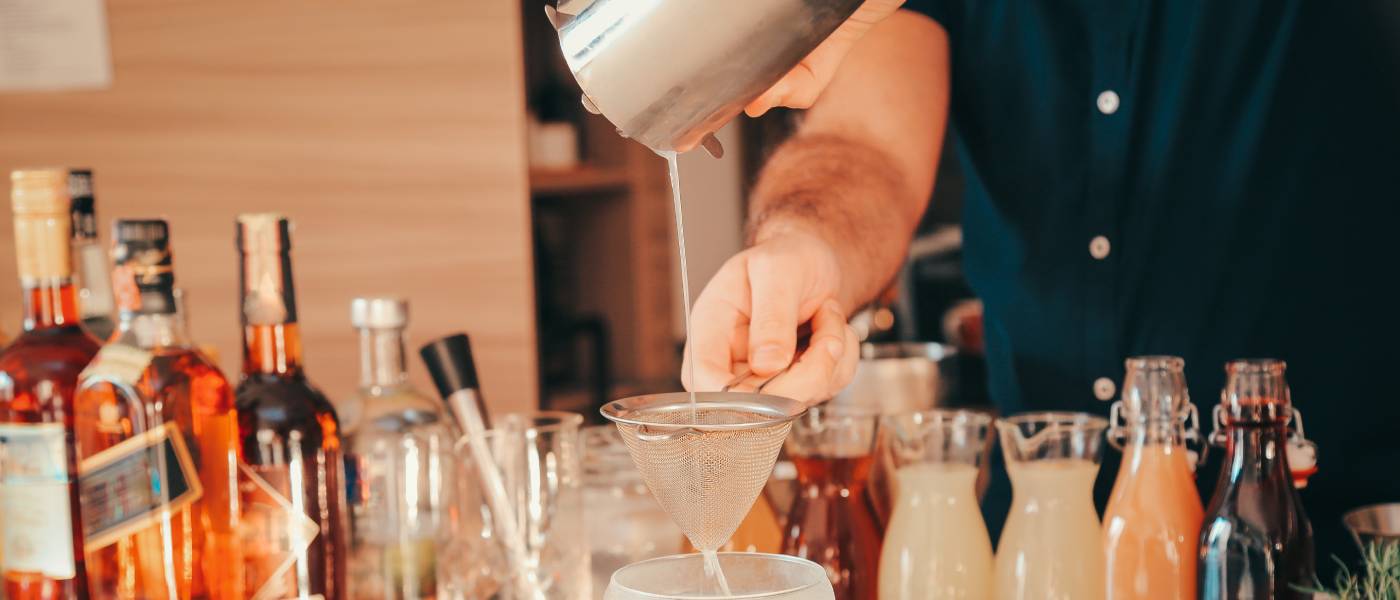 A man standing behind a home bar countertop, straining a drink into a glass. Several liquors and syrups line the counter.