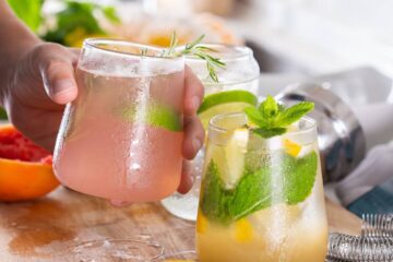 A man's hand holding a cocktail glass over a cutting board with other cocktails and several fruits scattered around.