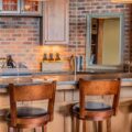 A Tuscan-inspired home bar with a dark stone countertop, faux brick wall, and three wood bar stools.