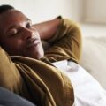 A young Black man with his hands behind his head and eyes closed lies back on a beige couch in a bright room.