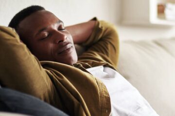 A young Black man with his hands behind his head and eyes closed lies back on a beige couch in a bright room.