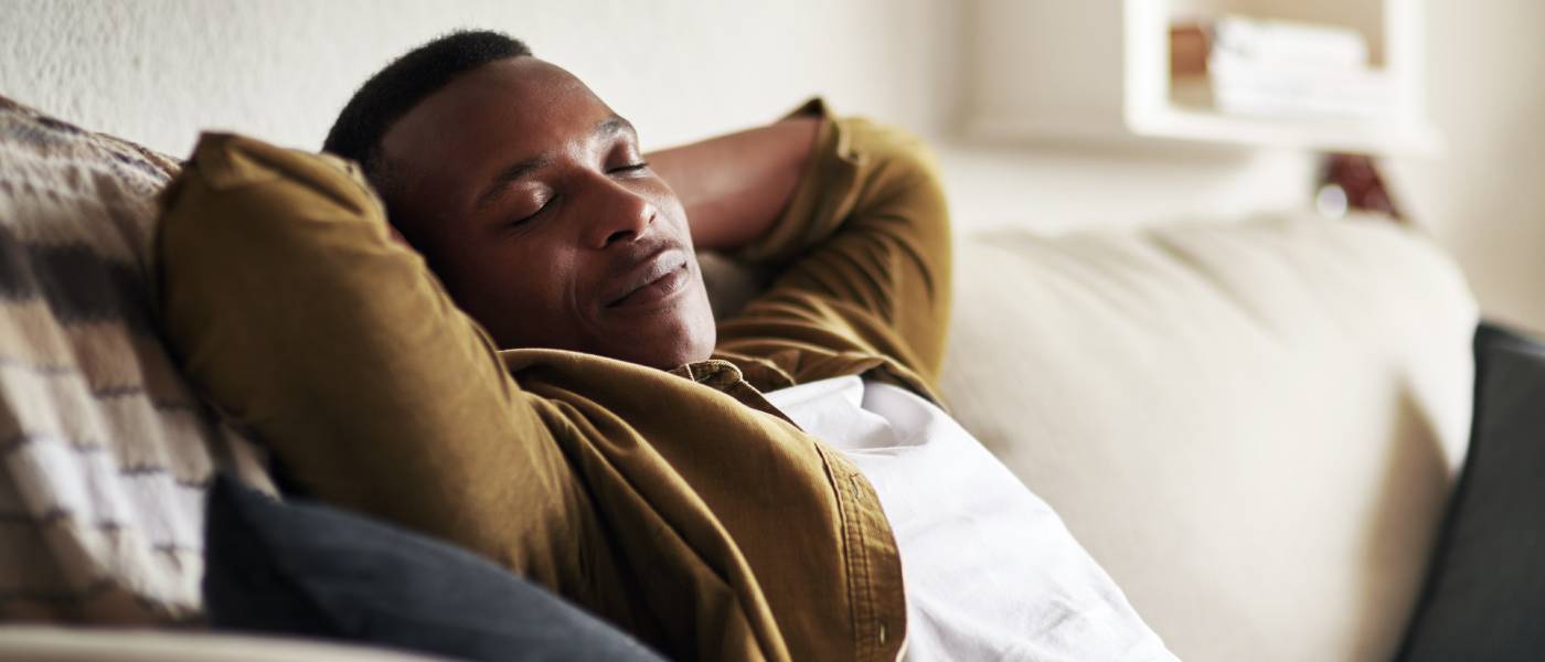 A young Black man with his hands behind his head and eyes closed lies back on a beige couch in a bright room.