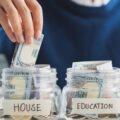 Four money-filled glass jars with the labels "Car," "House," "Education," "Investment." A blurred man is placing money into one.