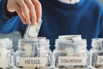 Four money-filled glass jars with the labels "Car," "House," "Education," "Investment." A blurred man is placing money into one.