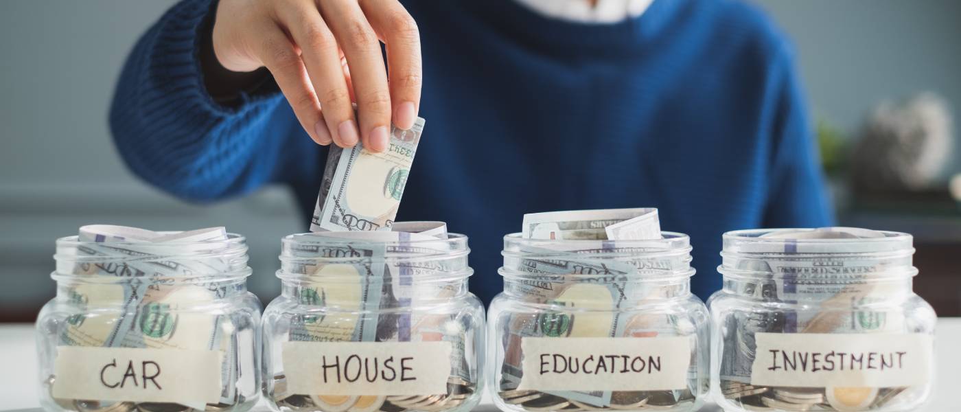 Four money-filled glass jars with the labels "Car," "House," "Education," "Investment." A blurred man is placing money into one.