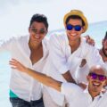 A group of men on the beach posing for a photo. They're smiling and wearing hats and sunglasses with their arms stretched.