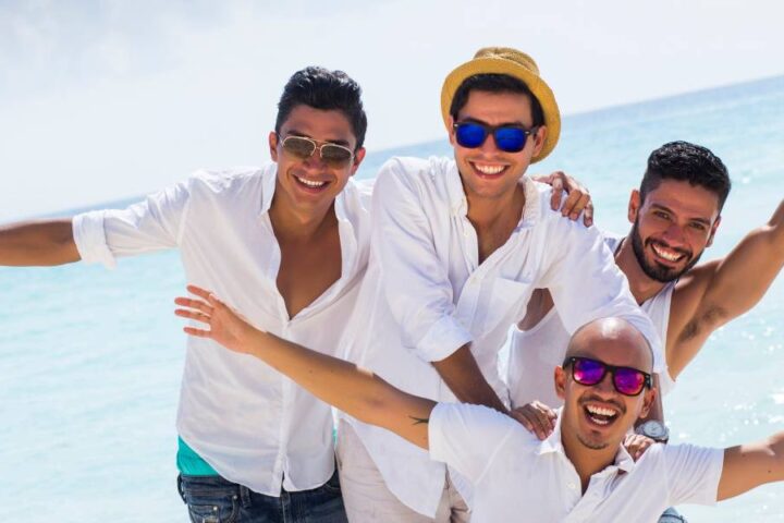 A group of men on the beach posing for a photo. They're smiling and wearing hats and sunglasses with their arms stretched.