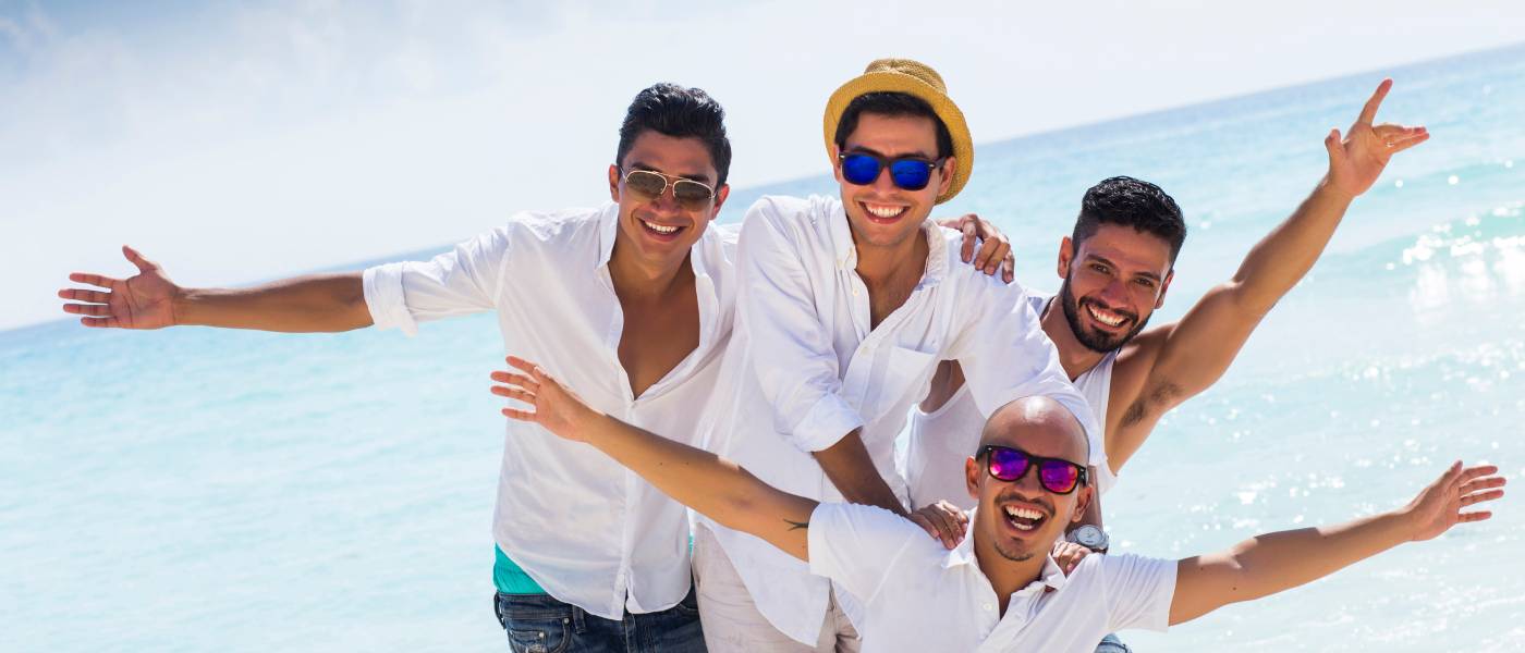 A group of men on the beach posing for a photo. They're smiling and wearing hats and sunglasses with their arms stretched.