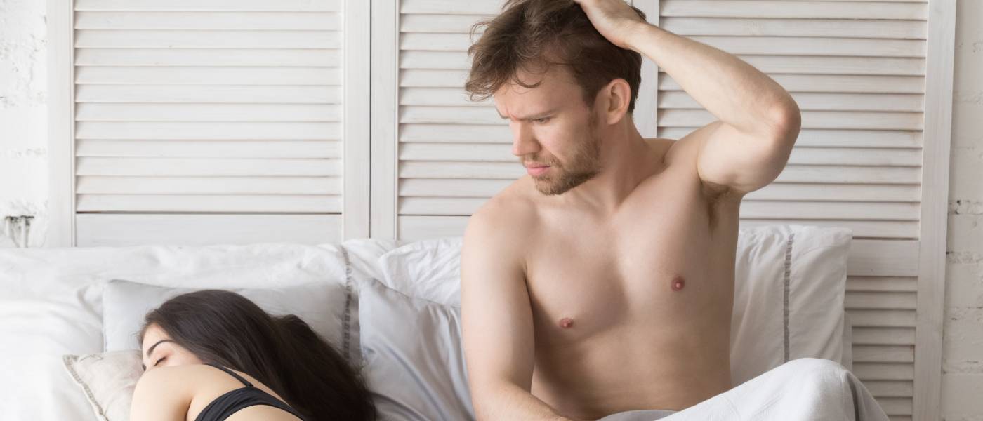 A couple in bed. The woman is sleeping, turned away from the man, and he is sitting up, tousling his hair and looking confused.