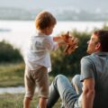 A man sits on the grass in front of a lake, looking up at a little boy playing with a wooden airplane.