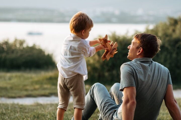 A man sits on the grass in front of a lake, looking up at a little boy playing with a wooden airplane.