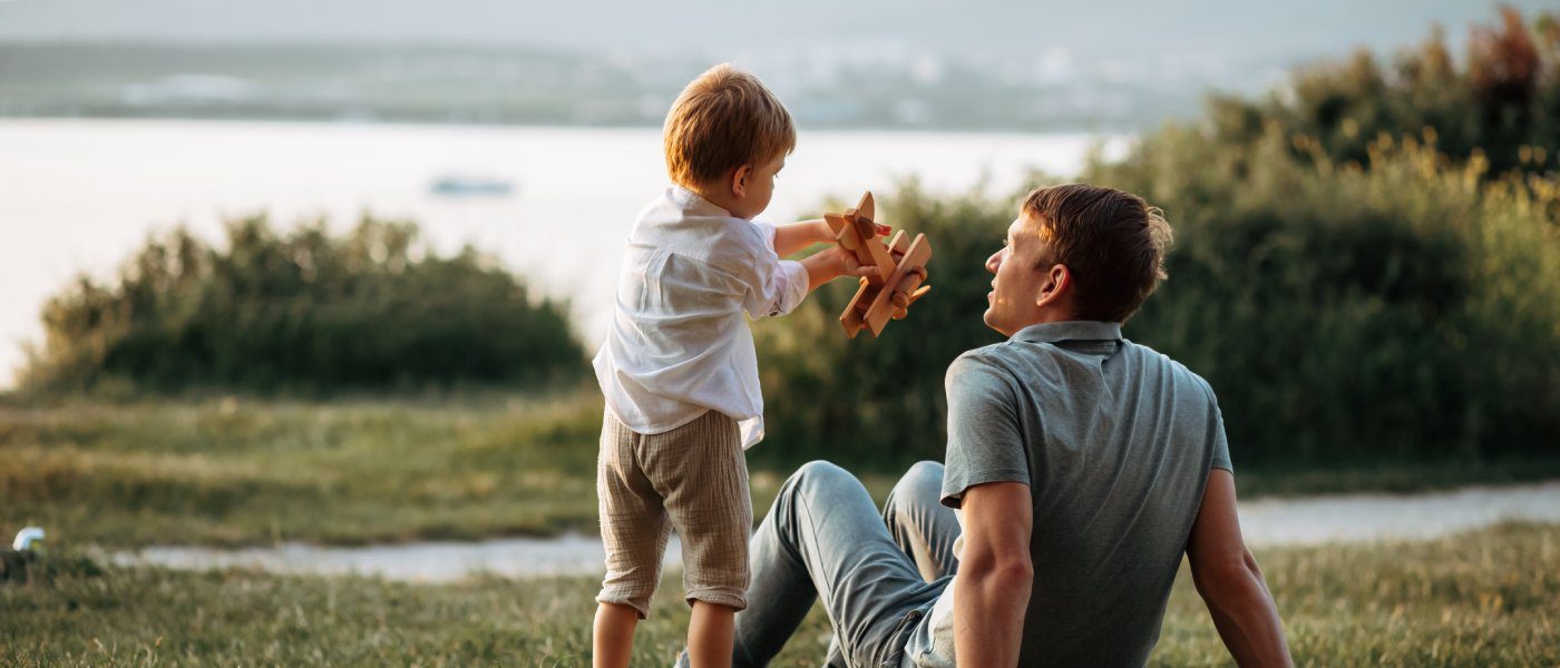 A man sits on the grass in front of a lake, looking up at a little boy playing with a wooden airplane.