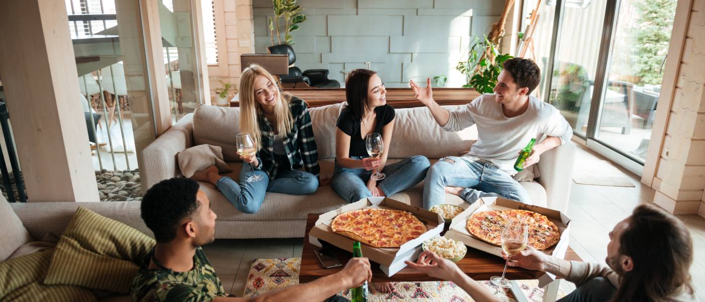 A group of friends sit around a small coffee table in a living room. They are sharing drinks, pizzas, and popcorn.