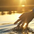 A person in a jacket riding on a still lake in a kayak. They are dragging their fingers along the surface of the water.