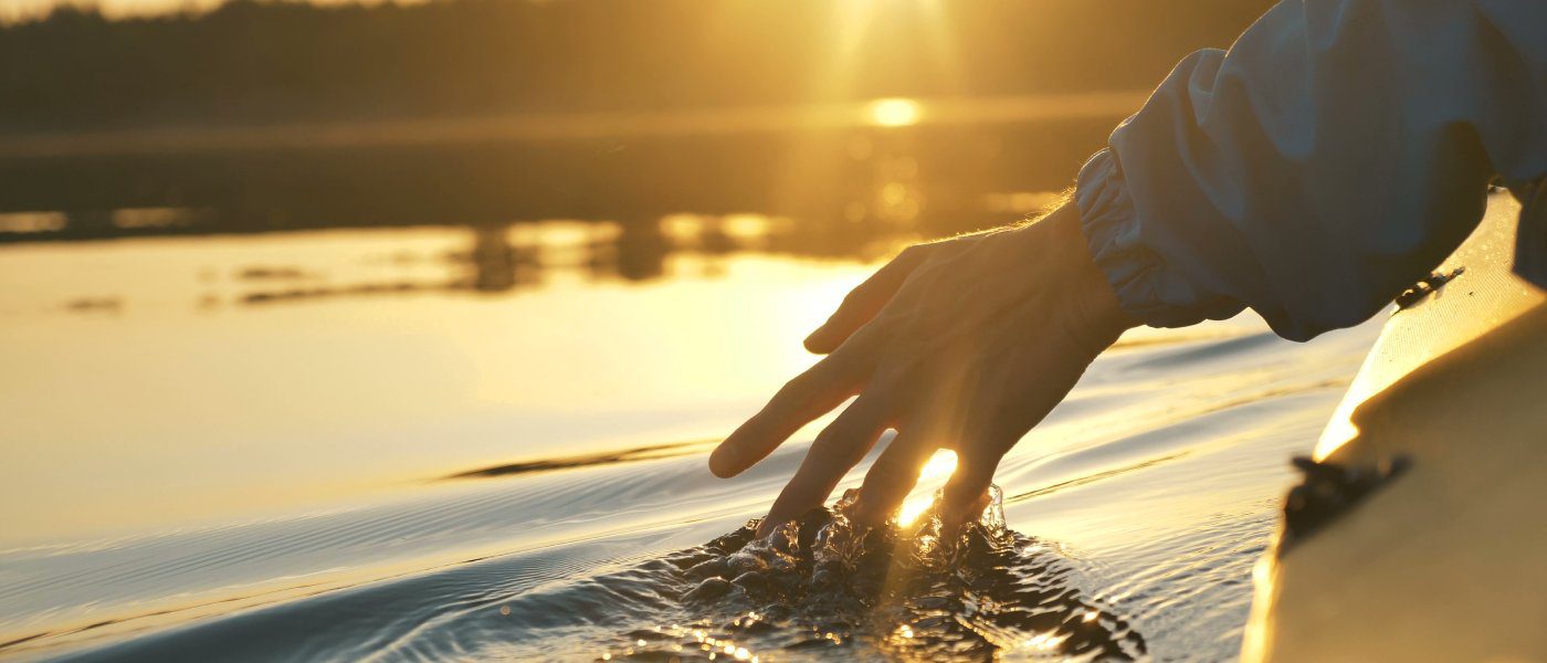 A person in a jacket riding on a still lake in a kayak. They are dragging their fingers along the surface of the water.