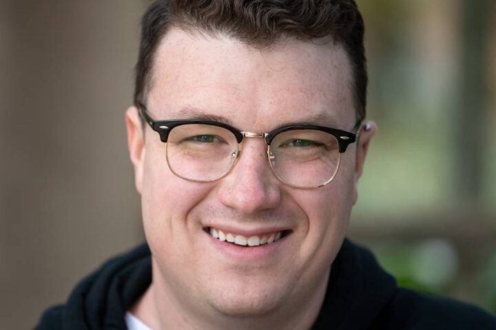 A professional headshot featuring a smiling young man with semi-rimless glasses and a neat haircut.