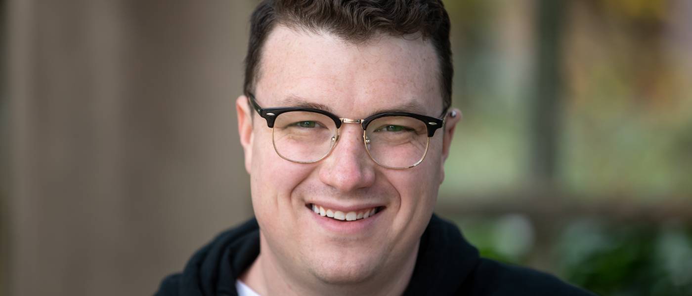 A professional headshot featuring a smiling young man with semi-rimless glasses and a neat haircut.