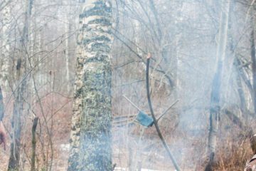 Two men in heavy hunting gear resting beside a small campfire and arranging sticks to hold a pot above the fire.