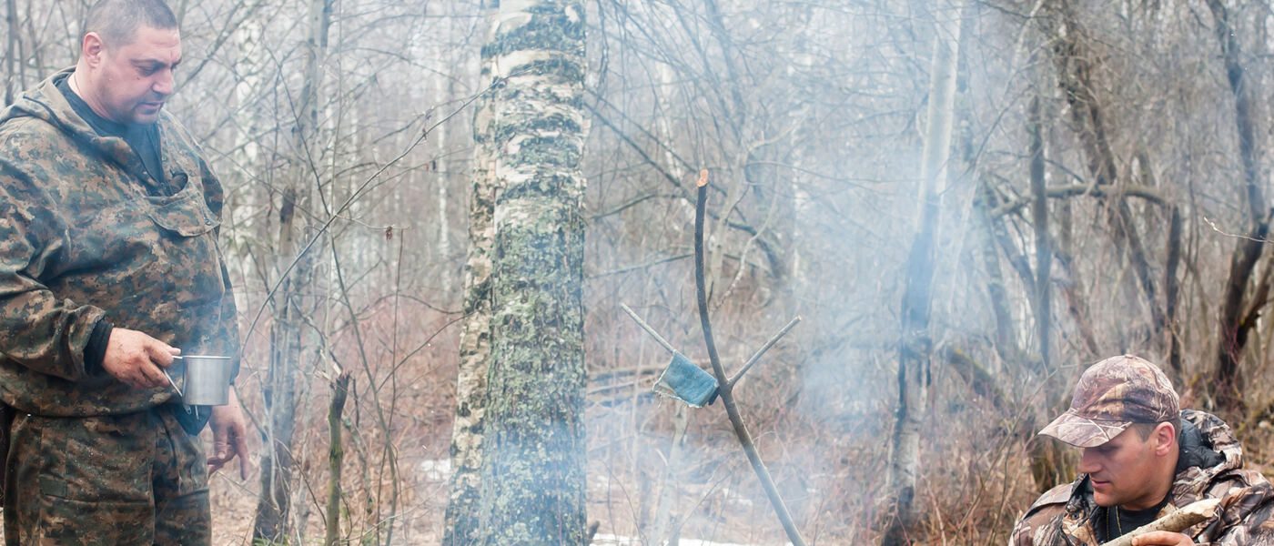 Two men in heavy hunting gear resting beside a small campfire and arranging sticks to hold a pot above the fire.