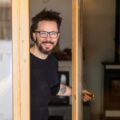 A man with glasses and tattoos opening the glass door to his well-kept home and giving the viewer a welcoming smile.