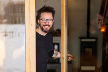 A man with glasses and tattoos opening the glass door to his well-kept home and giving the viewer a welcoming smile.