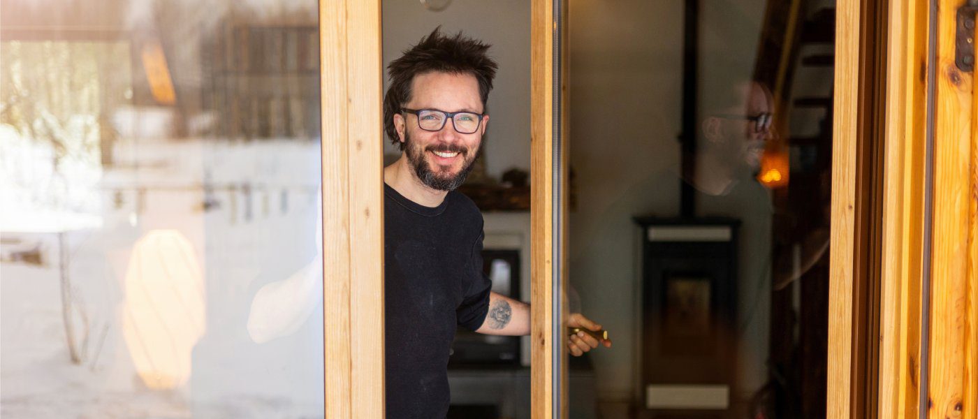 A man with glasses and tattoos opening the glass door to his well-kept home and giving the viewer a welcoming smile.