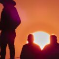 A shadow of two people in cowboy hats at sunset with a group beside them. The sky is orange and pink.