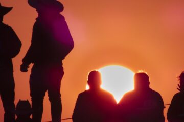 A shadow of two people in cowboy hats at sunset with a group beside them. The sky is orange and pink.