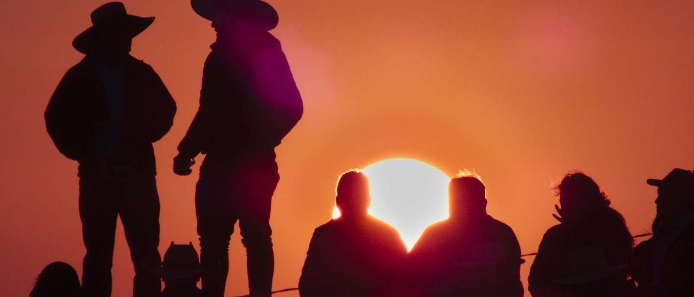 A shadow of two people in cowboy hats at sunset with a group beside them. The sky is orange and pink.