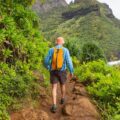 The backside of someone in a long-sleeved shirt, shorts, and a backpack hiking along a lush, dirt trail with the ocean below.