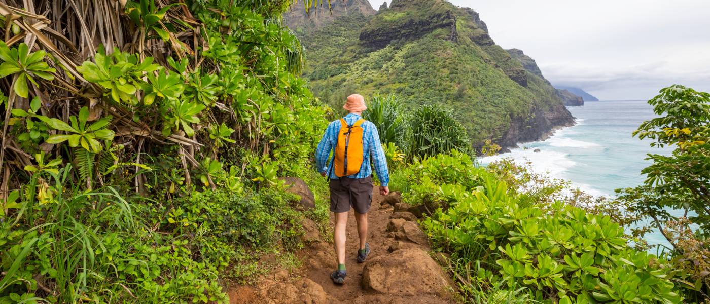 The backside of someone in a long-sleeved shirt, shorts, and a backpack hiking along a lush, dirt trail with the ocean below.