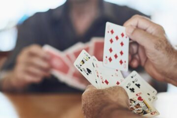 Two men sitting at a wooden table with multiple playing cards in their hands. Each card is of a different suit.