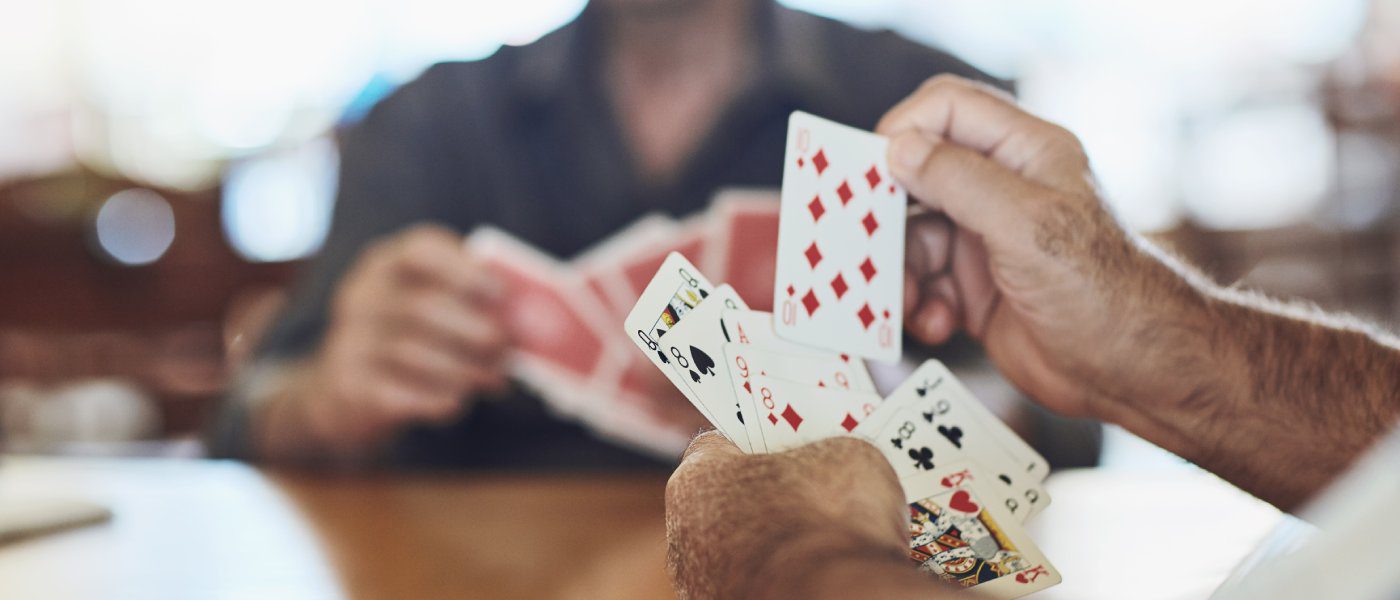 Two men sitting at a wooden table with multiple playing cards in their hands. Each card is of a different suit.