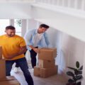 Two smiling men bringing several sealed cardboard moving boxes into the foyer of a white-and-beige home.