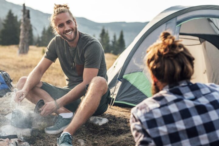 Two men sit by their campfire and tents as they cook food in a small metal pot, smiling and conversing.