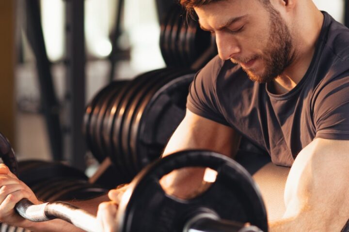 A concentrated young brunette man using a barbell to perform bicep curls over a curl bench in a gym.