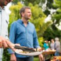 A man in a pale blue button-down shirt is pulling a kebab off a grill and putting it on a plate. Another man walks up to him.