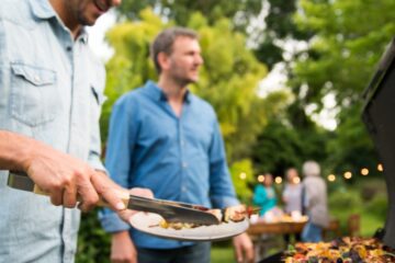 A man in a pale blue button-down shirt is pulling a kebab off a grill and putting it on a plate. Another man walks up to him.