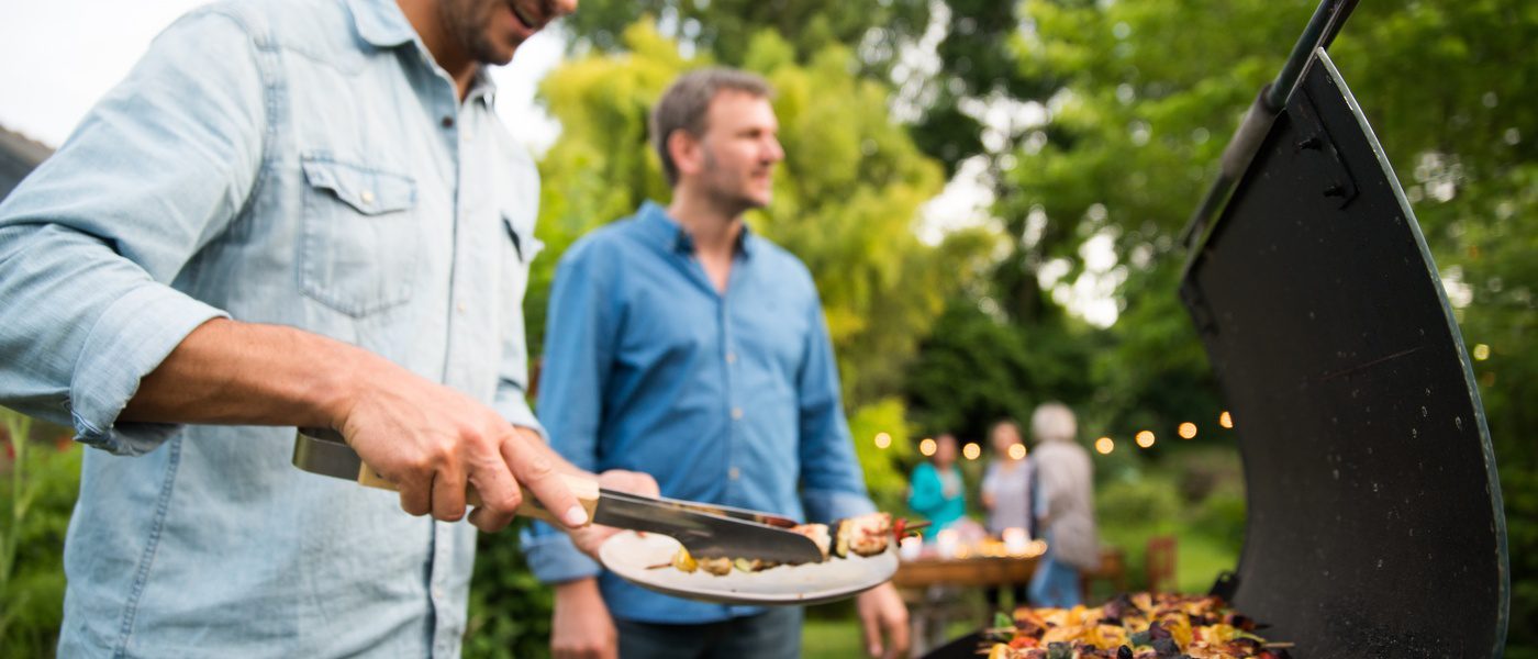 A man in a pale blue button-down shirt is pulling a kebab off a grill and putting it on a plate. Another man walks up to him.