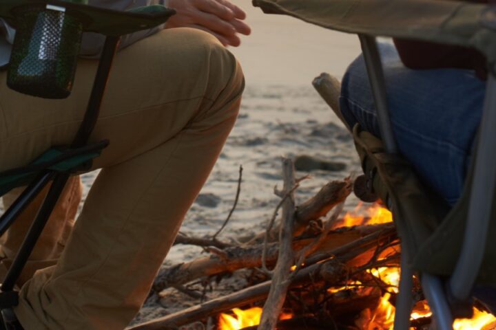 Two men are sitting in lawn chairs on a beach, a fire burning in front of them. The man on the left is turned to the other.