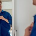 A young red-haired man grinning as he looks at himself in the mirror and buttons up his bright-blue dress shirt.