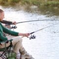 Two young men sit in camp chairs as they fish on a dock next to a river. One of them is reeling in a catch.