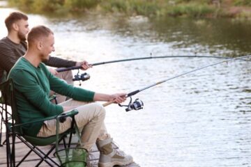 Two young men sit in camp chairs as they fish on a dock next to a river. One of them is reeling in a catch.