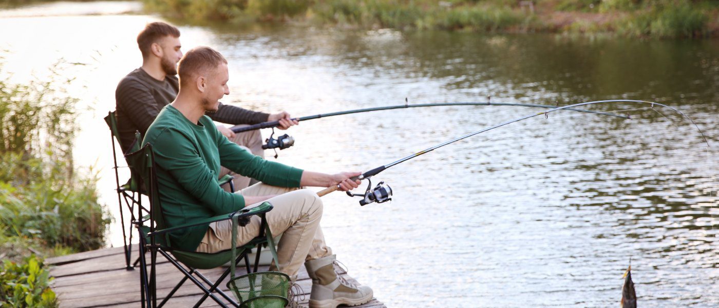 Two young men sit in camp chairs as they fish on a dock next to a river. One of them is reeling in a catch.